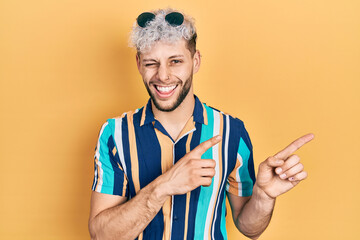 Young hispanic man with modern dyed hair pointing with fingers to the side winking looking at the...