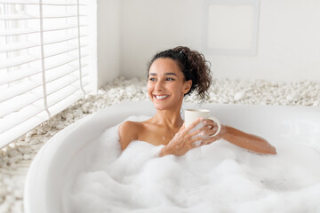 Naklejka premium Carefree young woman sitting in foamy bath, drinking coffee or tea near window at home, free space