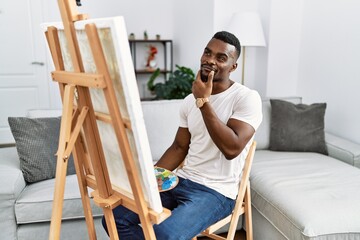 Young african man painting on canvas at home looking confident at the camera smiling with crossed arms and hand raised on chin. thinking positive.