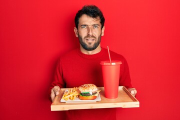 Handsome man with beard eating a tasty classic burger with fries and soda clueless and confused expression. doubt concept.