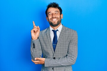 Handsome man with beard wearing business suit and tie with a big smile on face, pointing with hand and finger to the side looking at the camera.