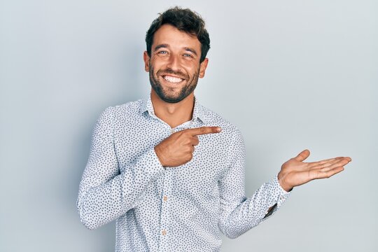 Handsome man with beard wearing casual elegant shirt amazed and smiling to the camera while presenting with hand and pointing with finger.