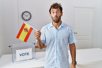 Young handsome man at political campaign election holding spain flag scared and amazed with open mouth for surprise, disbelief face