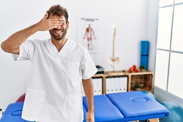 Young handsome physiotherapist man working at pain recovery clinic smiling and laughing with hand on face covering eyes for surprise. blind concept.