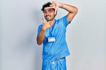 Young hispanic man wearing blue male nurse uniform smiling making frame with hands and fingers with...