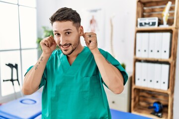 Young physiotherapist man working at pain recovery clinic smiling pulling ears with fingers, funny gesture. audition problem