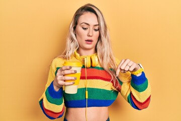 Beautiful young blonde woman drinking cup of coffee wearing headphones pointing down with fingers showing advertisement, surprised face and open mouth