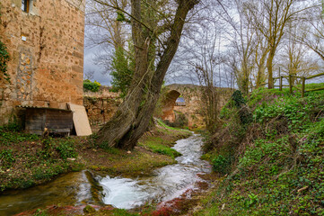 Fototapeta na wymiar close-up of a small waterfall in a stream that runs between the grass, trees, the wall of a medieval church and that in the background runs under a stone bridge from the Middle Ages the sky with dark 