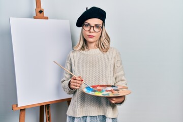 Beautiful caucasian woman drawing with palette on easel stand relaxed with serious expression on...