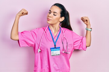 Young brunette woman wearing doctor uniform and stethoscope showing arms muscles smiling proud....