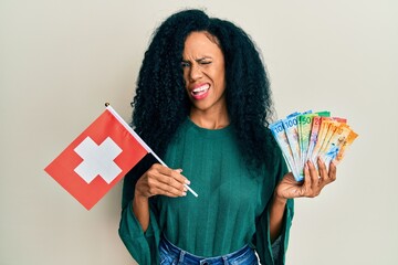 Middle age african american woman holding switzerland flag and franc banknotes winking looking at the camera with sexy expression, cheerful and happy face.