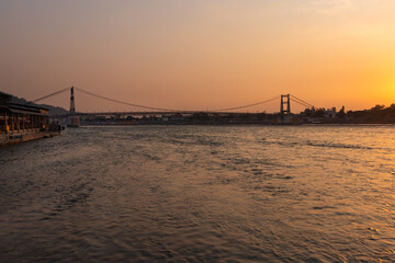 Fototapeta na wymiar iron suspension bridge with sunset orange sky over flowing river horizon at evening from flat angle