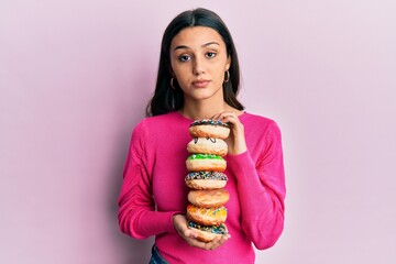 Young hispanic woman holding pile of tasty colorful doughnuts relaxed with serious expression on...