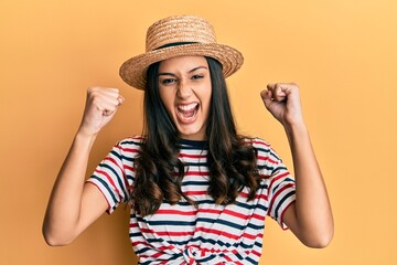 Young hispanic woman wearing summer hat screaming proud, celebrating victory and success very excited with raised arms