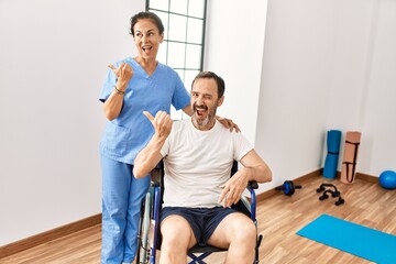 Obraz premium Hispanic middle age man sitting on wheelchair and nurse at rehabilitation clinic pointing thumb up to the side smiling happy with open mouth