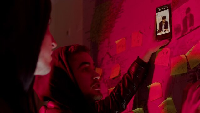 Two young men standing in front of wall map with strings, stickers and photos and comparing image to the one on smartphone, talking and pointing. Side view. Investigation, cyber crime concept