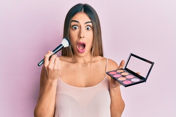 Young hispanic woman holding makeup brush and blush afraid and shocked with surprise and amazed...