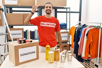 Young hispanic man wearing volunteer t shirt at donations stand smiling looking to the camera...