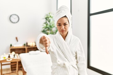 Young brunette woman wearing towel and bathrobe standing at beauty center looking unhappy and angry showing rejection and negative with thumbs down gesture. bad expression.