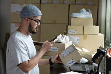 Asian male small business owner using mobile app on smartphone checking parcel box. Warehouse worker, seller holding phone scanning retail drop shipping package postal parcel bar code. - 499591648