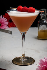 A French Martini cocktail garnished with fresh raspberries on a white background and styled with bar equipment and flowers.