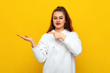 Beautiful young brunette woman in white casual style sweatshirt indicating fingers on palm empty...
