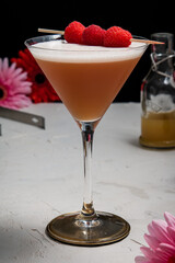 A French Martini cocktail garnished with fresh raspberries on a white background and styled with bar equipment and flowers.