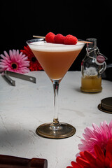 A French Martini cocktail garnished with fresh raspberries on a white background and styled with bar equipment and flowers.