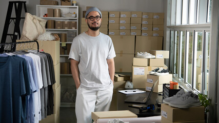 Confident young Asian man retail seller, entrepreneur, online store drop shipping small business owner looking at camera standing in delivery shipping warehouse with parcel boxes.