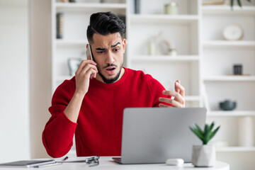 Upset attractive millennial arabic middle eastern in red clothes call by phone, gesticulate, look at laptop in home