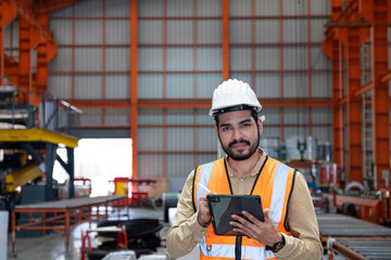 Selective focus at Asian men engineer supervisor, wearing safety equipment. While using digital tablet for quality and safety control inside of factory area. With blurred background of heavy machine.