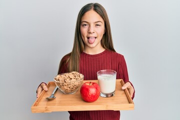 Beautiful brunette little girl holding tray with breakfast food sticking tongue out happy with funny expression.