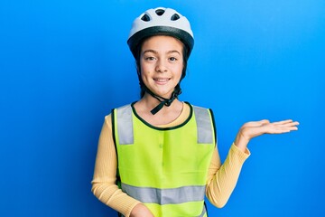 Beautiful brunette little girl wearing bike helmet and reflective vest smiling cheerful presenting and pointing with palm of hand looking at the camera.