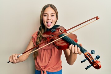 Beautiful brunette little girl playing violin sticking tongue out happy with funny expression.