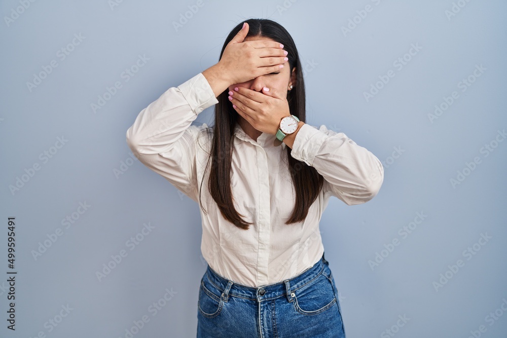 Sticker Young latin woman standing over blue background covering eyes and mouth with hands, surprised and shocked. hiding emotion