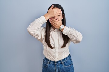 Fototapeta premium Young latin woman standing over blue background covering eyes and mouth with hands, surprised and shocked. hiding emotion