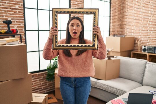 Young latin woman moving to a new home holding empty frame afraid and shocked with surprise and amazed expression, fear and excited face.