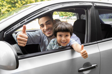 Glad happy millennial arab guy teaches little boy to drive car, dad and son show thumbs up in open...