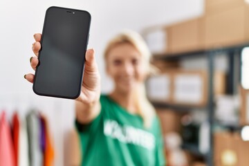Young blonde volunteer girl smiling happy showing app smartphone at charity center.
