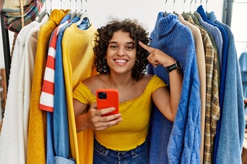 Young hispanic woman searching clothes on clothing rack using smartphone smiling pointing to head with one finger, great idea or thought, good memory