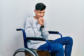 Young hispanic man sitting on wheelchair laughing nervous and excited with hands on chin looking to...