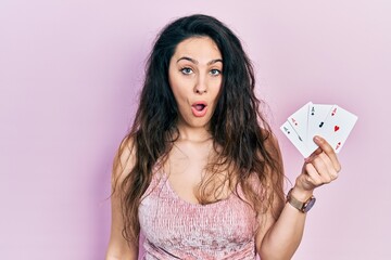 Young hispanic woman holding poker cards scared and amazed with open mouth for surprise, disbelief face