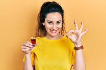 Young hispanic woman drinking whiskey shot doing ok sign with fingers, smiling friendly gesturing excellent symbol