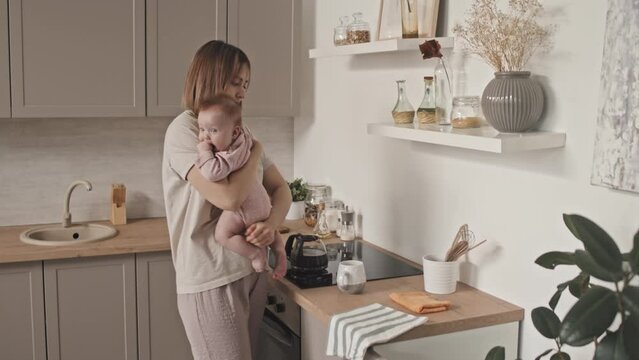 Medium long of young busy Biracial woman in loungewear holding adorable infant, standing in kitchen at daytime, wiping kitchen table and electric burner