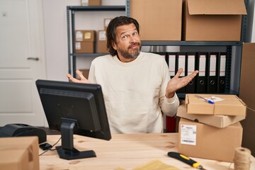 Handsome middle age man working at small business ecommerce clueless and confused expression with arms and hands raised. doubt concept.