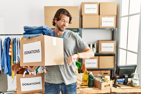 Handsome middle age man holding donations box for charity at volunteer stand checking the time on wrist watch, relaxed and confident