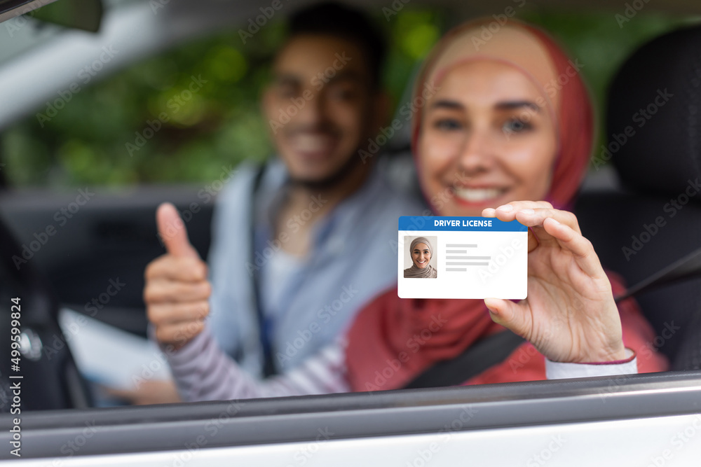 Wall mural Glad middle eastern male and female in hijab show driving license and thumb up in open window in car after exam