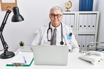 Senior caucasian man wearing doctor uniform and stethoscope at the clinic winking looking at the...