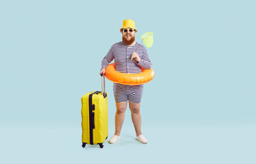 Full body length of happy funny fat man in striped swim suit, hat, glasses and floaty standing in studio and holding yellow suitcase and butterfly scoop net. Traveling vacation, summer holiday concept