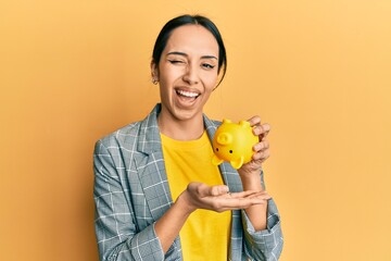 Young hispanic girl holding piggy bank winking looking at the camera with sexy expression, cheerful...